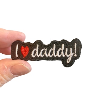 Può includere: Un cerotto in feltro nero con ricamo bianco che dice "Ti amo papà!" con un cuore rosso.