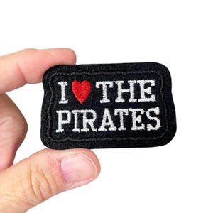 May include: Black rectangular patch with white embroidered text "I ❤️ THE PIRATES" and a red heart. The patch has a black border and is held by a hand against a white background. The patch is a fan item.