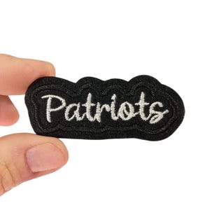 May include: A black embroidered patch with the word "Patriots" in white script. The patch has a cloud-like border and is held between two fingers.