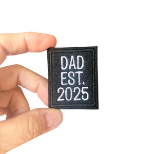 以下が含まれることがあります： 「DAD EST. 2025」と白い刺繍が施された黒い長方形のパッチ。白い背景にパッチが掲げられています。