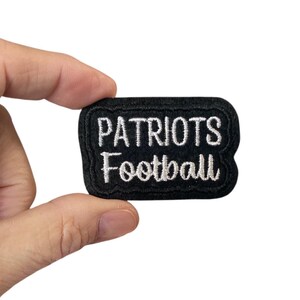 May include: Black embroidered patch with the words "PATRIOTS Football" in white lettering. The patch has a black border and is held between fingers against a white background. A sports-themed item.
