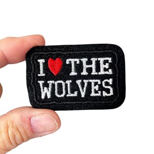 May include: A black rectangular patch with white embroidered text that reads "I ❤️ THE WOLVES." The heart is red. The patch has a black border.