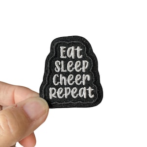 May include: A black embroidered patch featuring the text "Eat Sleep Cheer Repeat" in white. The patch has a wavy edge and is held up by a hand. The patch is likely for a cheerleader and is against a white background.