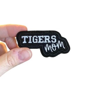 Op de afbeelding: Zwart geborduurde patch met de witte woorden "TIGERS mom". De patch heeft een zwarte rand en wordt door een hand tegen een witte achtergrond gehouden. De patch heeft een rechthoekige vorm met een gebogen rand.