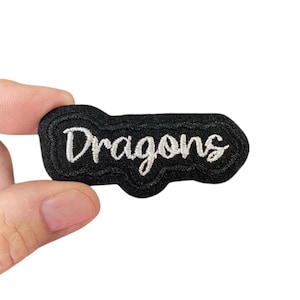 Może przedstawiać: Czarna łatka z filcu z białym haftem z napisem "Dragons".