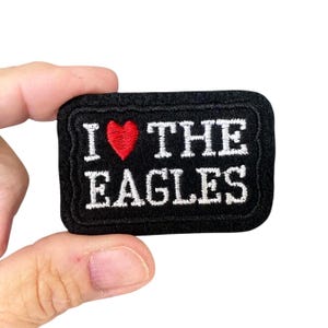 May include: Black rectangular patch with the text "I ❤️ THE EAGLES" embroidered in white. A red heart replaces the word "love". The patch has a black border.