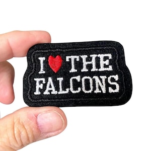 May include: Black rectangular patch with the text "I ❤️ THE FALCONS" embroidered in white. A red heart replaces the word "love". The patch has a black border and is held by a hand.