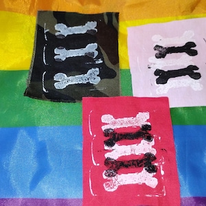 May include: Three rectangular fabric patches featuring bone-shaped prints. One patch is camouflage with three white bone prints, one is light pink with two black bone prints, and one is red with four alternating black and white bone prints.