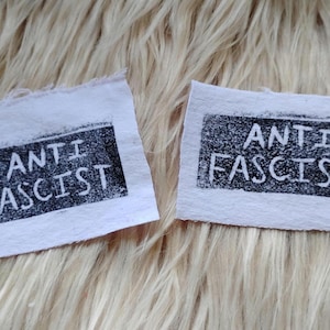 May include: Two rectangular white fabric patches with raw, frayed edges. Each patch features a black rectangular block print containing the white text ANTI FASCIST in a bold, sans-serif, hand-stamped style. The patches are separate and distinct.