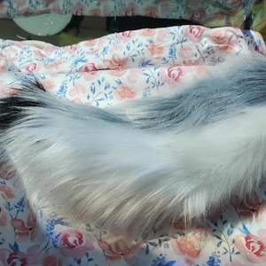 May include: A fluffy, faux fur tail in shades of white, grey, and black. The tail is lying on a floral patterned fabric with pink and blue flowers. The tail is a cosplay or costume accessory.