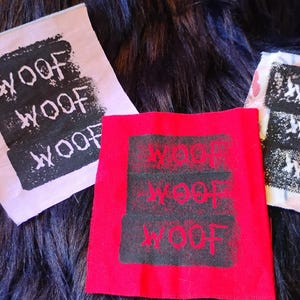 May include: Three square fabric patches featuring the text WOOF WOOF WOOF printed in a distressed black font. One patch is light pink, one is bright red, and the third is a white fabric with a subtle floral pattern in soft pastel colors.