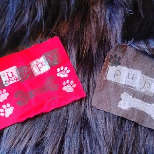 May include: Two rectangular fabric patches, one red and one dark brown, each featuring the word puppy printed in white block letters. Both patches display a white bone graphic and several white paw prints arranged around the text.