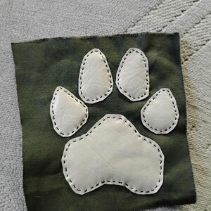 May include: A square patch featuring a white paw print design with black stitching, set against a dark green fabric background. The paw print is composed of five individual pads. The patch is likely for decorative purposes.