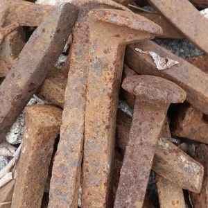 Puede incluir: Primer plano de varios clavos de ferrocarril grandes y oxidados. Los clavos tienen un vástago rectangular y una cabeza ancha y plana. El metal está muy corroído, con un color marrón rojizo, lo que indica edad y exposición. Los clavos están dispersos.