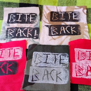 May include: Five rectangular fabric patches featuring the text BITE BACK in a distressed, stencil-style font. The patches come in various colors including white with floral patterns, beige, red, and camouflage, each with contrasting ink colors.