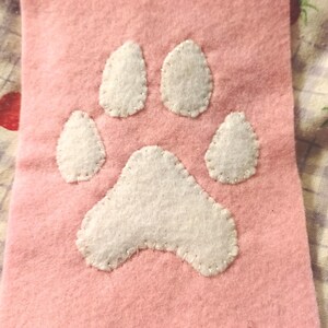 May include: A pink felt square with a white paw print stitched on it. The paw print is made of white felt and has five toes. The background is a fabric with a strawberry pattern.