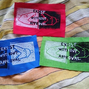 May include: Three rectangular fabric patches in red, blue, and green. Each patch features a screen-printed graphic of a fish-like creature split between two contrasting color blocks, with the text "EXIST WITHOUT APPROVAL" printed in white.