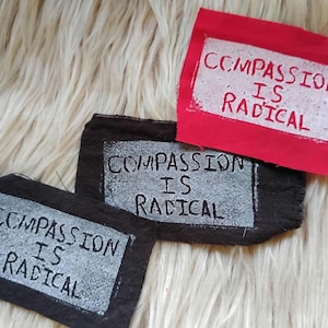 May include: Three rectangular fabric patches featuring the text COMPASSION IS RADICAL printed in a distressed style. One patch is bright red with white text, while the other two are black with white text, all showing raw, unfinished edges.