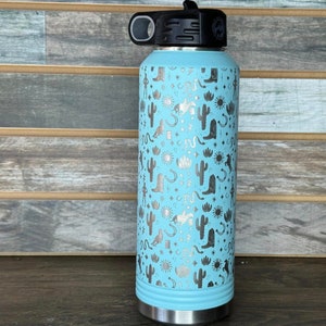 May include: A light blue stainless steel water bottle with a silver design of cacti, boots, and stars. The bottle has a black lid with a loop handle.