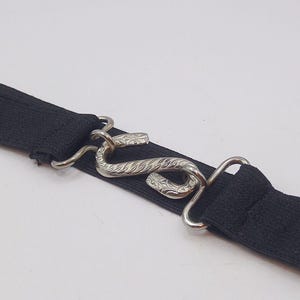 May include: A close-up shot of a black elasticated belt featuring a silver snake-shaped clasp. The clasp has detailed engraved patterns. The belt is designed for waist wear.