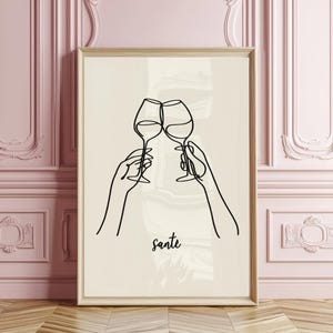 Peut inclure: Une affiche encadrée avec un dessin au trait minimaliste de deux verres de vin qui s'entrechoquent, tenus par des mains. Le mot "sante" est écrit en cursive en dessous des verres. L'œuvre est sur fond crème, encadrée de bois clair.
