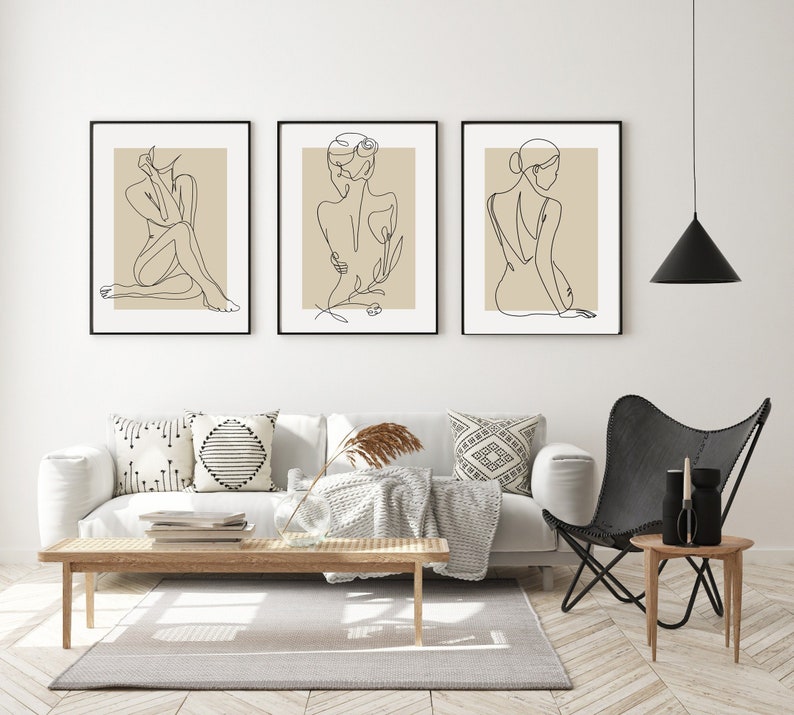 Puede incluir: Tres impresiones minimalistas de arte lineal con figuras femeninas y detalles florales. Las impresiones est&aacute;n enmarcadas en negro y se muestran en una pared blanca sobre un sof&aacute; blanco con almohadas y una manta.