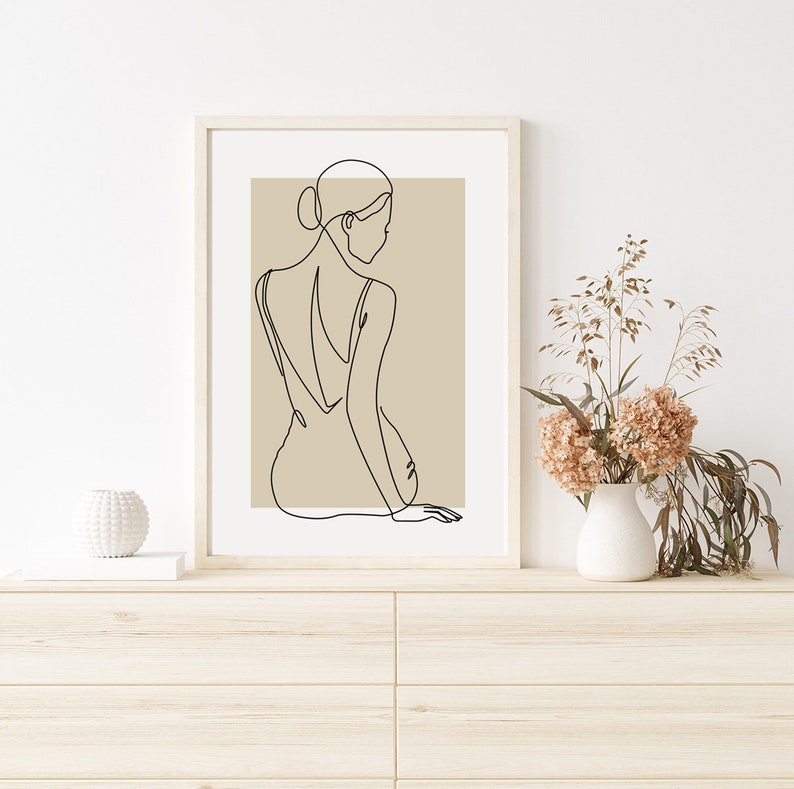 Puede incluir: Una impresi&oacute;n enmarcada de arte lineal de una mujer sentada de espaldas al espectador. El fondo de la impresi&oacute;n es de color marr&oacute;n claro. La impresi&oacute;n est&aacute; colgada en una pared blanca sobre una c&oacute;moda de madera.