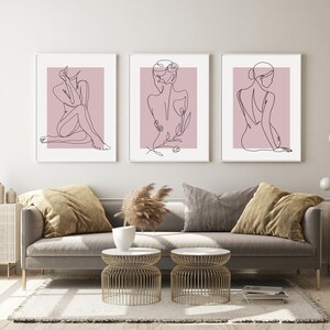 Pu&ograve; includere: Tre stampe minimaliste di arte lineare di figure su uno sfondo rosa chiaro. Le stampe sono incorniciate in bianco e appese a una parete bianca sopra un divano grigio.