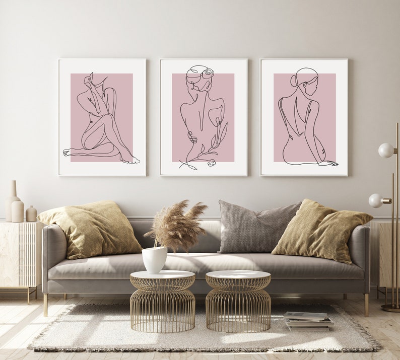 Pu&ograve; includere: Tre stampe minimaliste di arte lineare di figure su uno sfondo rosa chiaro. Le stampe sono incorniciate in bianco e appese a una parete bianca sopra un divano grigio.