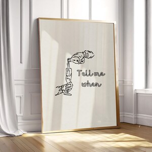 Peut inclure: Une affiche encadrée avec une illustration en noir et blanc d'une main versant d'une bouteille dans une flûte à champagne. Le texte "Tell me when" est écrit en cursive. Le cadre est doré.