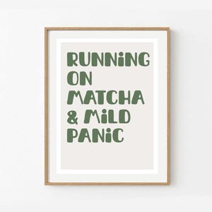 Könnte beinhalten: Gerahmter Kunstdruck mit dem Text "RUNNING ON MATCHA & MILD PANIC" in grünen, fetten Buchstaben. Der Druck hat einen hellbeigen Hintergrund und ist in einem Holzrahmen gefasst. Ein humorvolles Kunstwerk.