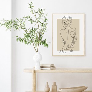 Puede incluir: Una impresi&oacute;n minimalista de arte de l&iacute;nea de una mujer con una flor detr&aacute;s de ella, enmarcada en un marco de madera claro y colgada en una pared blanca. La impresi&oacute;n est&aacute; sobre un fondo beige.
