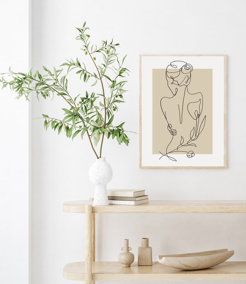 Puede incluir: Una impresi&oacute;n minimalista de arte de l&iacute;nea de una mujer con una flor detr&aacute;s de ella, enmarcada en un marco de madera claro y colgada en una pared blanca. La impresi&oacute;n est&aacute; sobre un fondo beige.