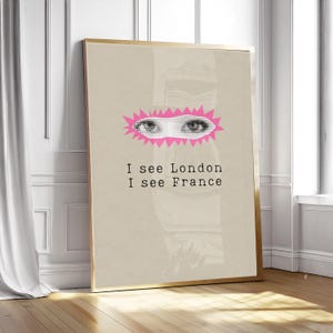Peut inclure: Une affiche encadrée avec un cadre doré, représentant un design d'yeux roses et blancs et le texte "I see London I see France". L'œuvre est posée sur un fond beige, contre un mur blanc et un sol en bois.