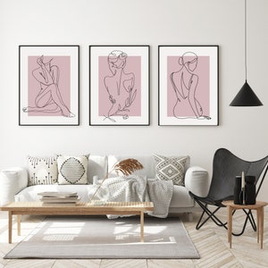Pu&ograve; includere: Tre stampe minimaliste di arte lineare di una persona in varie pose su uno sfondo rosa. Le stampe sono incorniciate in nero e appese a un muro bianco.