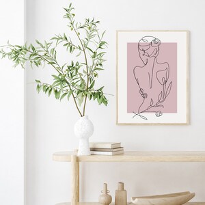 Pu&ograve; includere: Un disegno a linea di una persona con un fiore dietro, stampato su uno sfondo rosa. La stampa &egrave; incorniciata e appesa a un muro bianco.