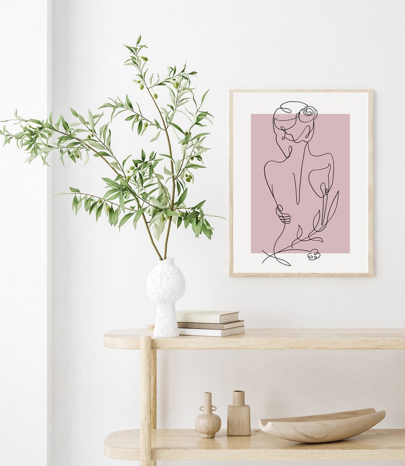 Pu&ograve; includere: Un disegno a linea di una persona con un fiore dietro, stampato su uno sfondo rosa. La stampa &egrave; incorniciata e appesa a un muro bianco.
