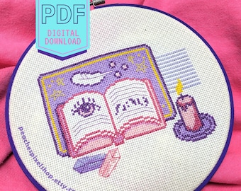 Puede incluir: Un patrón de punto de cruz morado y rosa de un libro abierto con una pluma, una vela y cristales. El libro tiene la palabra "FINIS" escrita en él. El patrón está en un aro de bordado redondo.