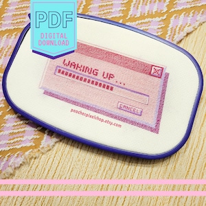 May include: A cross-stitch pattern of a computer screen with the text "Waking Up..." and a progress bar. The screen is pink and purple with a blue border. The pattern is on white Aida cloth and is framed in a blue hoop.