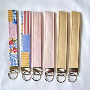 Könnte beinhalten: Sechs Schlüsselanhänger aus Stoff in verschiedenen Farben und Mustern. Die Schlüsselanhänger umfassen Blumen-, Streifen- und unifarbene Designs in Rosa, Gelb und Beige. Jeder Schlüsselanhänger hat einen silberfarbenen Metallring zur Befestigung von Schlüsseln.