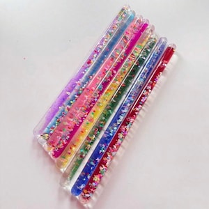 Puede incluir: Una colección de varitas de movimiento de líquido transparente y colorido. Cada varita está llena de purpurina y líquido en varios colores, incluyendo morado, rosa, verde, amarillo y azul. Las varitas están dispuestas en diagonal sobre una superficie blanca.