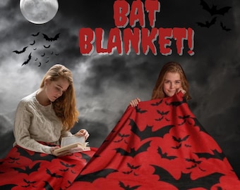 Può includere: Una coperta rossa con silhouette di pipistrelli neri, con la scritta "BAT BLANKET!". La coperta è mostrata con due persone, una che legge un libro e l'altra che tiene la coperta. Lo sfondo presenta una luna e pipistrelli.