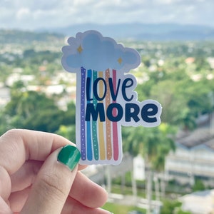 Peut inclure: Un autocollant avec un arc-en-ciel et un nuage avec les mots "Love More" en texte noir.