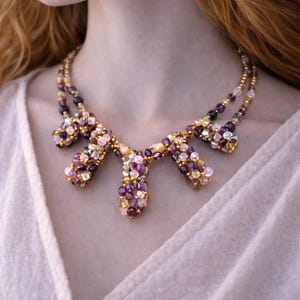 Peut inclure: Un collier de perles avec une chaîne dorée et des grappes de perles violettes, roses et blanches. Le collier est porté sur un haut blanc, créant un contraste avec les pierres précieuses colorées.