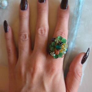 Peut inclure: Une main portant une bague ornée d'un groupe de perles vertes et d'accents colorés. Les ongles longs sont recouverts d'un vernis sombre et scintillant. La bague est le point central, présentant un design unique et accrocheur.