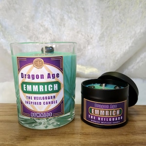 May include: Two candles in glass jars with black lids. The candles are teal blue and have a label that reads "Dragon Age Emmrich The Veilguard Inspired Candle Atmalis".