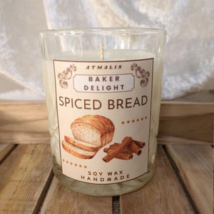Könnte beinhalten: Eine Glas-Kerze mit einem weißen Etikett, auf dem "Atmalis Baker Delight Spiced Bread" steht und eine Zeichnung von Brot und Zimtstangen abgebildet ist. Das Etikett trägt außerdem den Schriftzug "Soy Wax Handmade".