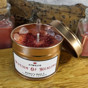 May include: A copper-colored candle tin with a red and glitter candle inside. The candle is labeled "Potion of Healing" with the text "Atmalis Baldur's Gate 3 Inspired Candle".