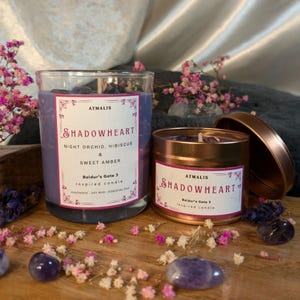 May include: Two candles with amethyst crystals inside. The candles are labeled "Shadowheart" and are inspired by the video game Baldur's Gate 3. The candles are in a glass jar and a tin container.