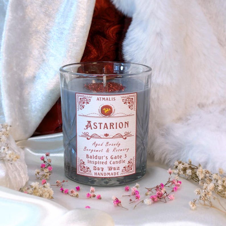 May include: A gray glass candle jar with a red and white label that reads "ATMALIS", "ASTARION", "Aged Brandy Bergamot & Rosemary", "Baldur's Gate 3 Inspired Candle", "Soy Wax", and "HANDMADE".